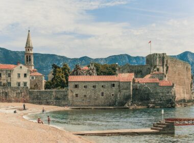 Unrecognizable tourists on sandy shore near ocean and aged church facades behind mounts under cloudy sky